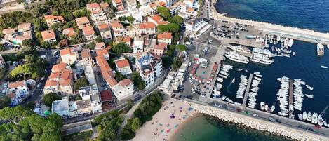Vue aérienne d’une ville côtière avec des immeubles résidentiels, une marina remplie de bateaux et une plage parsemée de parasols.