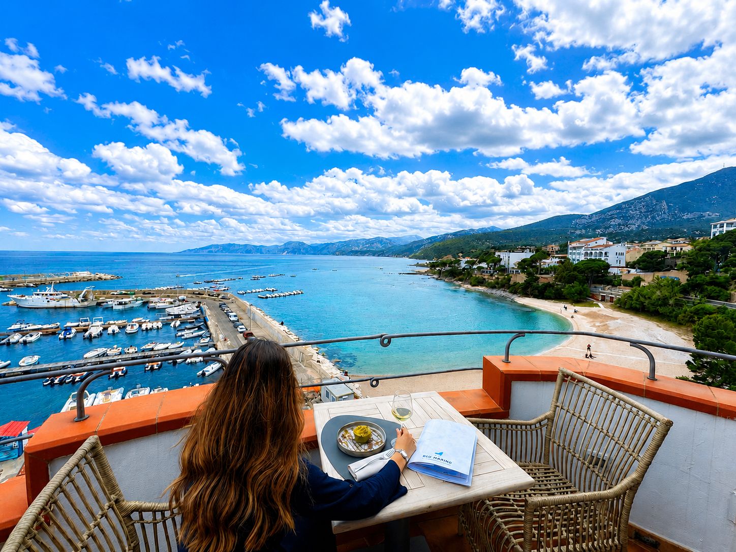 Un balcon avec une table et des chaises donnant sur une marina où sont amarrés des bateaux, ainsi que sur une plage.
