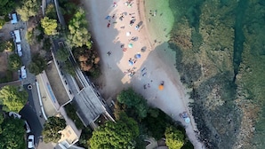 Vista aérea de una playa con agua cristalina, rodeada de vegetación y una carretera con vehículos.