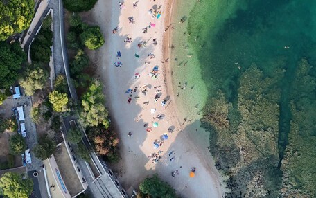 An aerial view of a beach with clear water, surrounded by greenery and a road with vehicles.