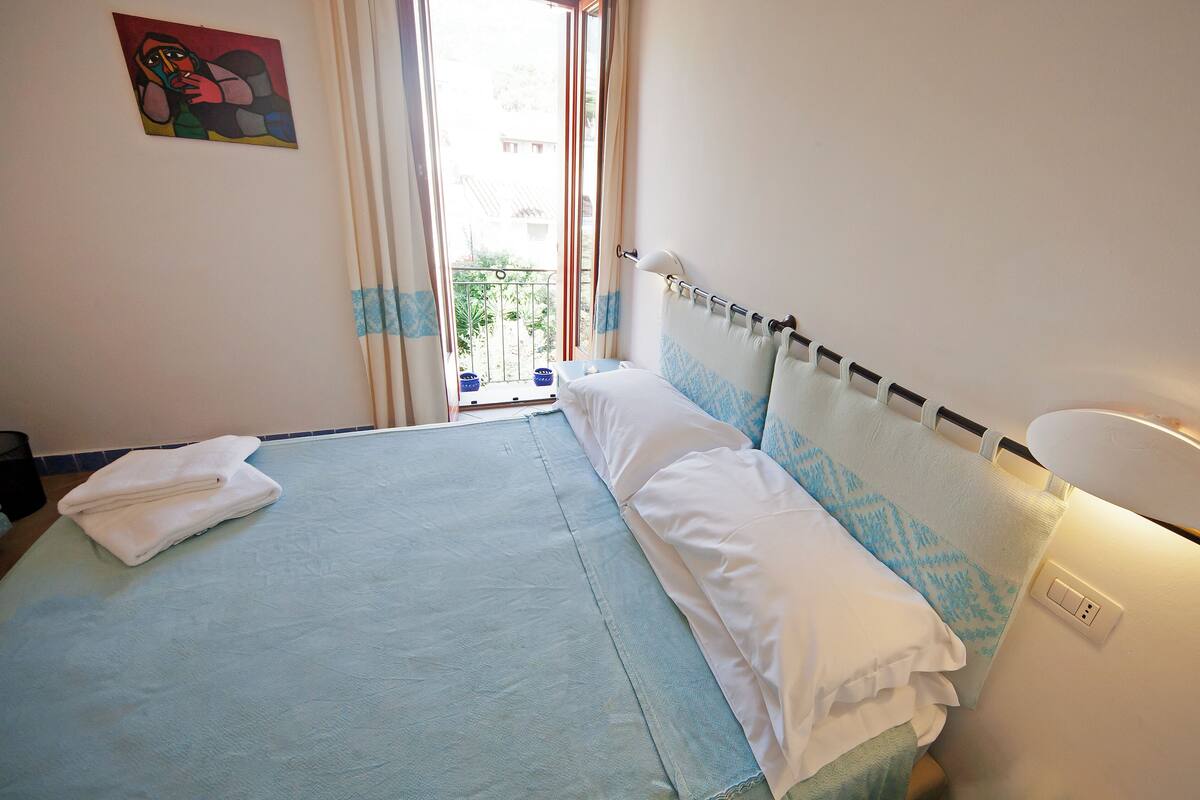 A hotel room with a bed, a window with curtains, a painting on the wall, and a bedside lamp.