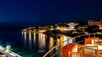 A nighttime view from a balcony overlooking a coastal area with a dining area featuring wooden furniture and a view of the sea.
