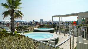 A rooftop pool area with lounge chairs, a palm tree, and a view of a beach with umbrellas. - Hotel Diamond (Riccione)