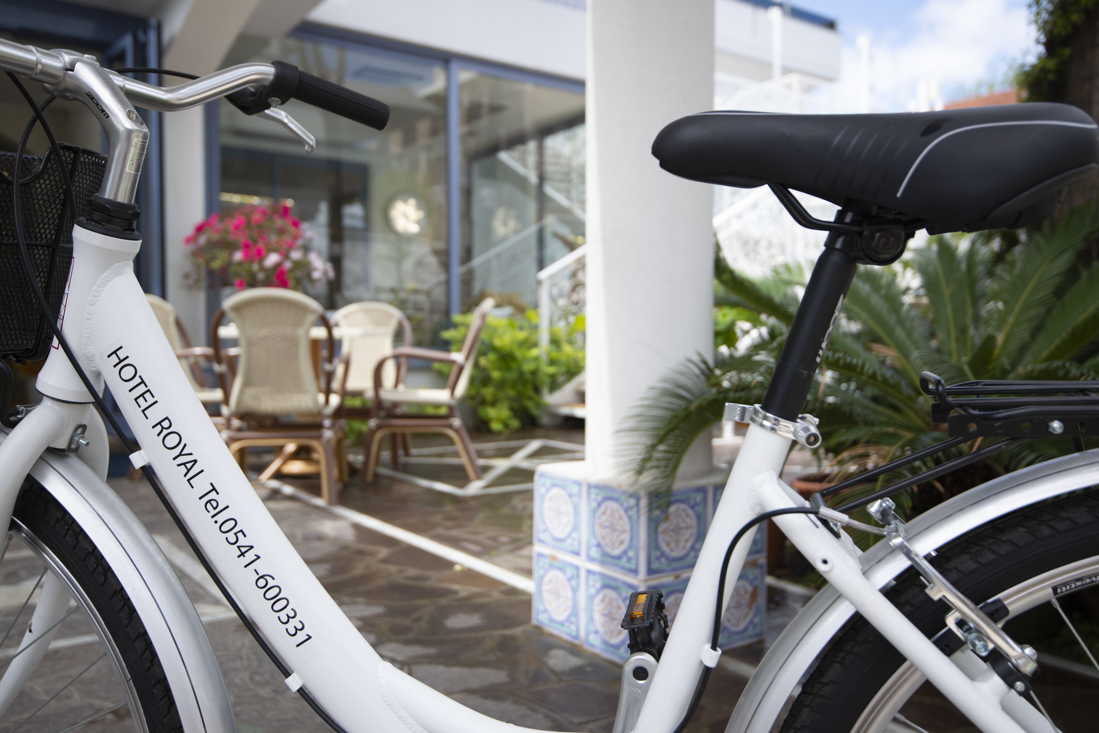 A white bicycle with a contact number, parked near a building entrance with outdoor seating and potted plants.