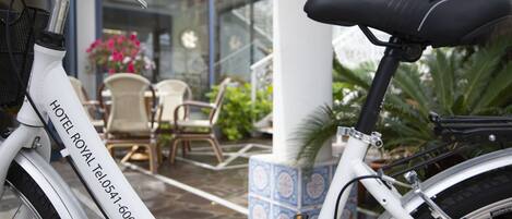 A white bicycle with a contact number, parked near a building entrance with outdoor seating and potted plants.
