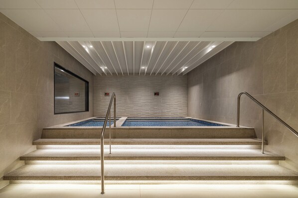 Un área moderna de piscina interior con vista despejada, escalones de acceso y una pared de azulejos con un reloj.