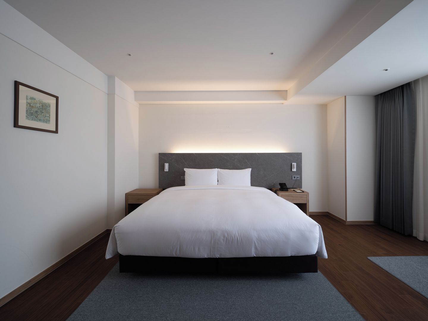 Ein modernes Hotelzimmer mit einem großen Bett, Nachttischen, einem Beistelltisch, einem gerahmten Bild an der Wand und einem Fenster mit Vorhängen.