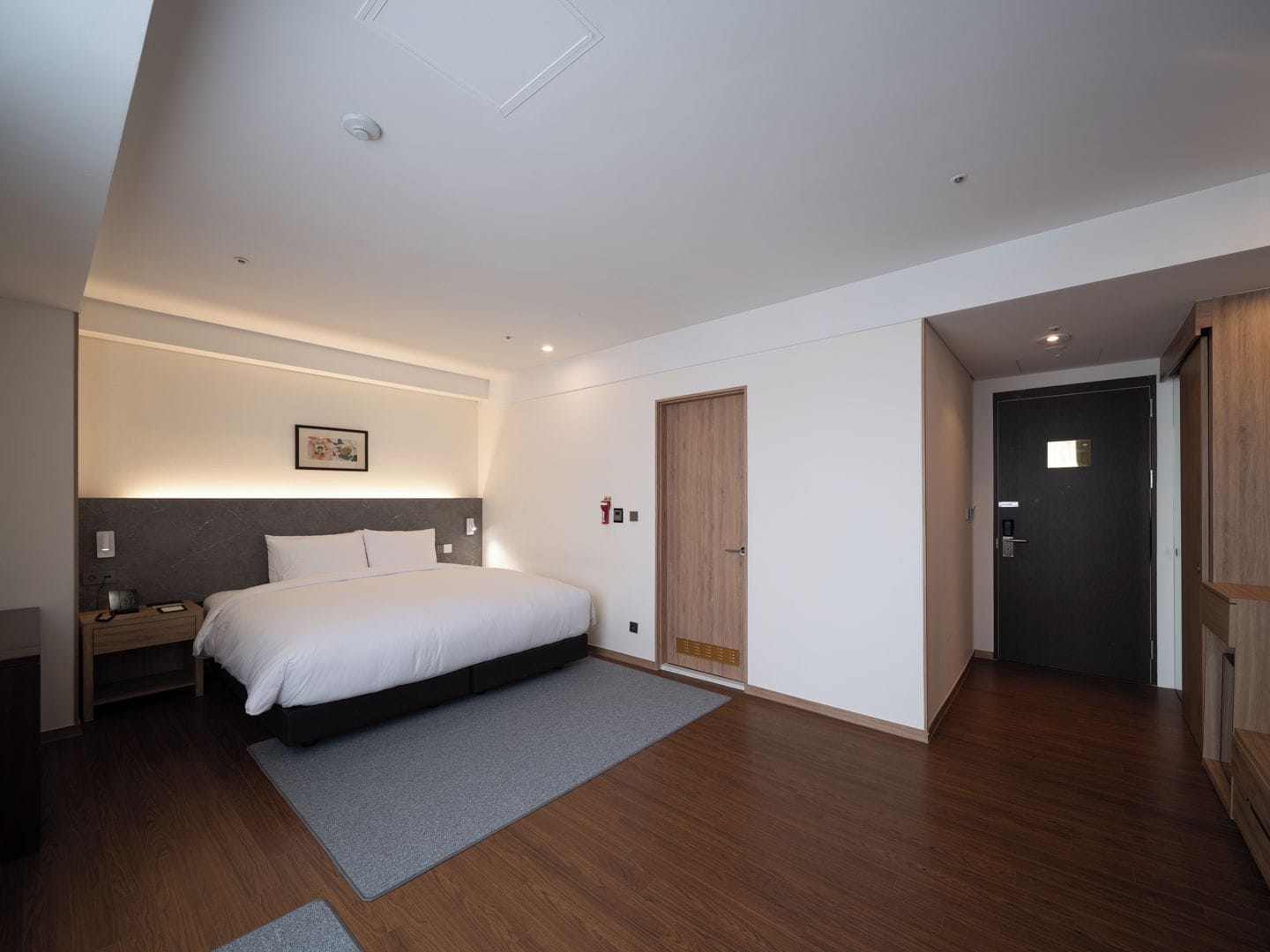 A modern hotel room with a large bed, a bedside table, a nightstand, a wardrobe, and a door leading to another room.