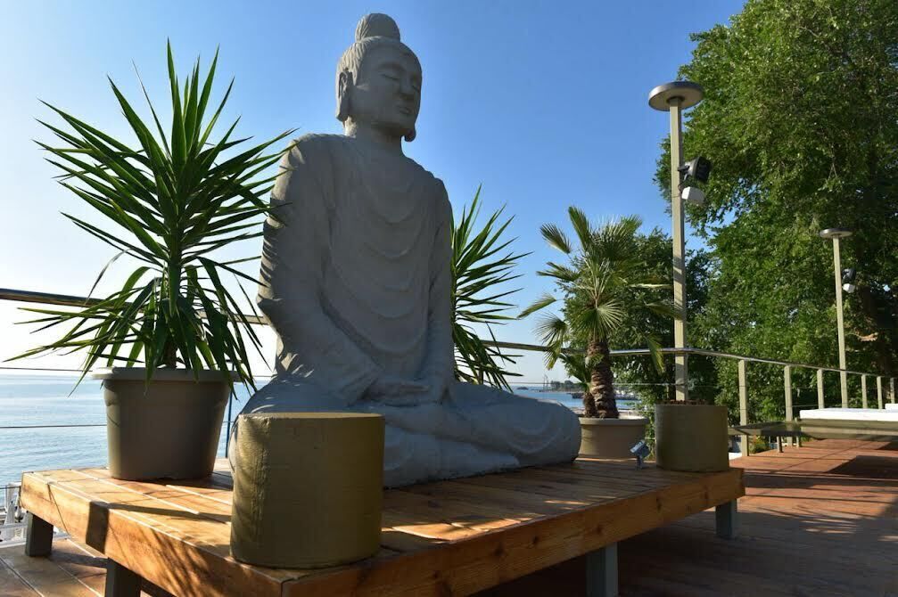 Eine sitzende Buddha-Statue auf einer Holzplattform mit Topfpflanzen, mit Blick auf ein Gewässer.