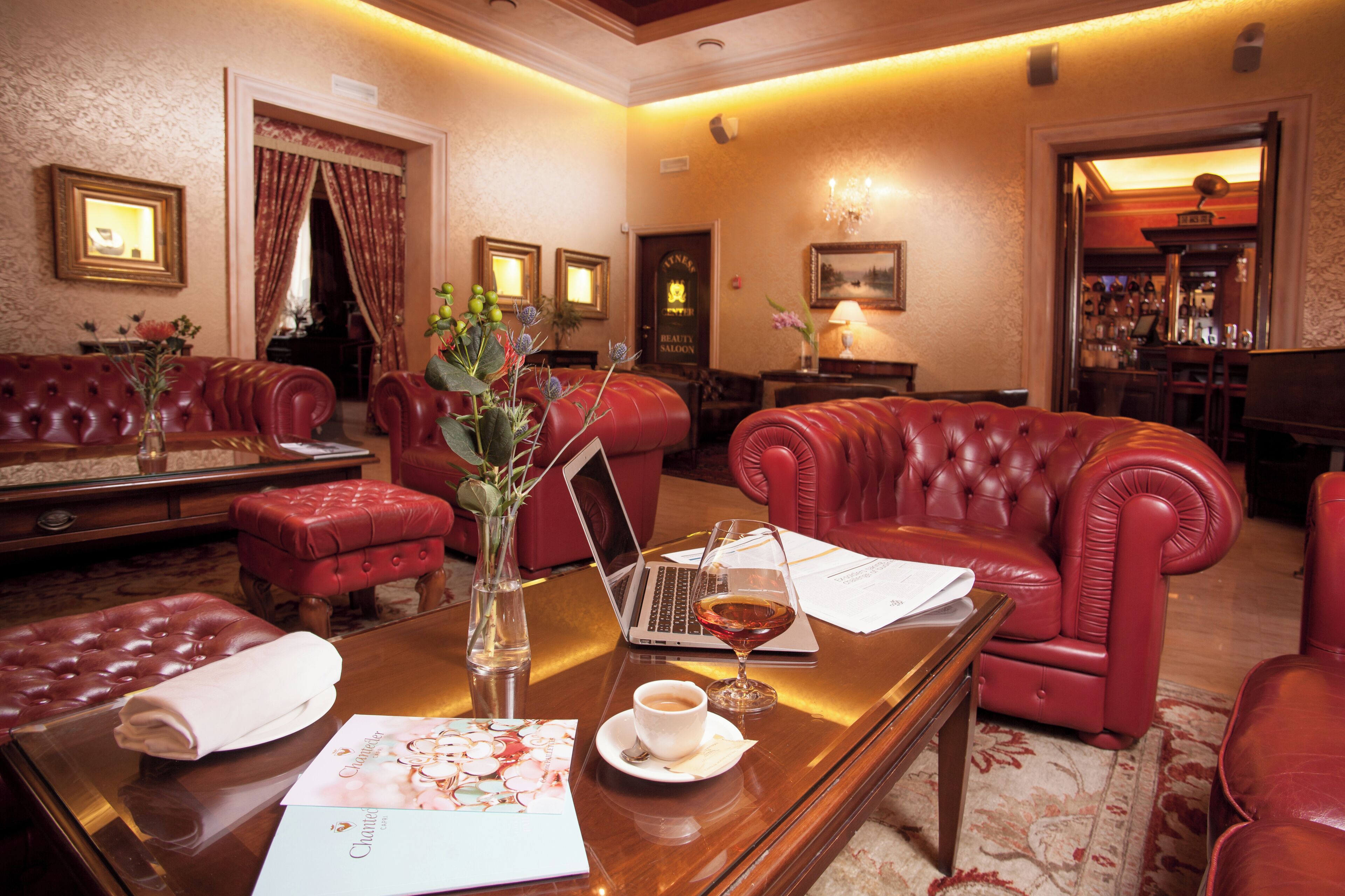 A hotel lobby with leather sofas, a wooden coffee table, a laptop, and a bar area in the background.