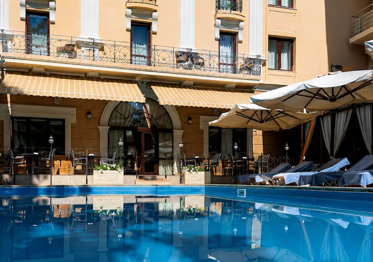 A hotel pool area with lounge chairs and umbrellas, a building with balconies, and a clear blue pool.