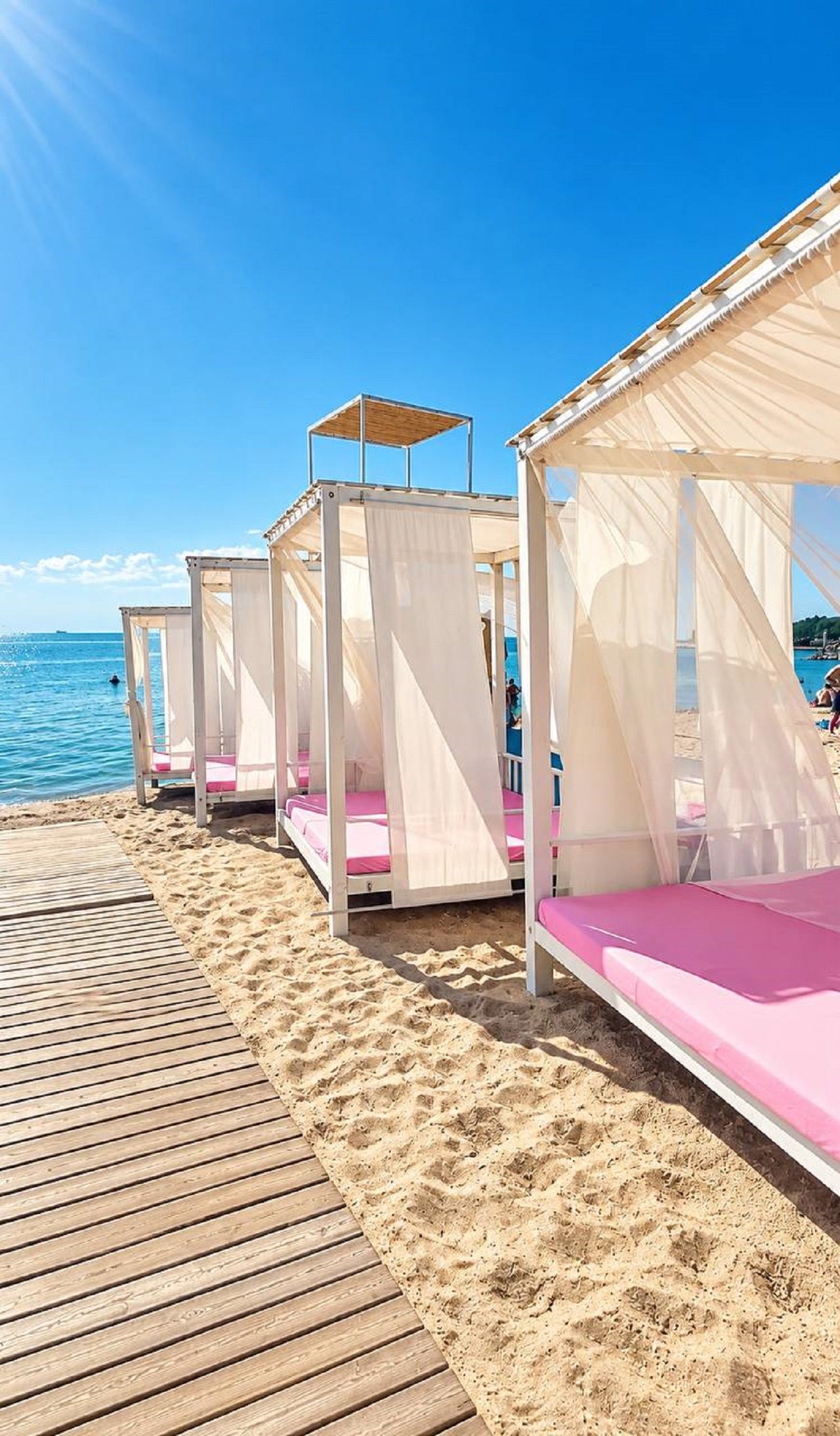 Eine Reihe von Strandkabinen mit weißen Markisen und rosafarbenen Kissen, auf einer Holzterrasse am Meer gelegen.
