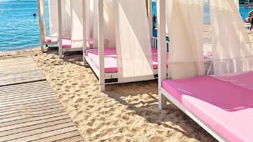 Eine Reihe von Strandkabinen mit weißen Markisen und rosafarbenen Kissen, auf einer Holzterrasse am Meer gelegen.