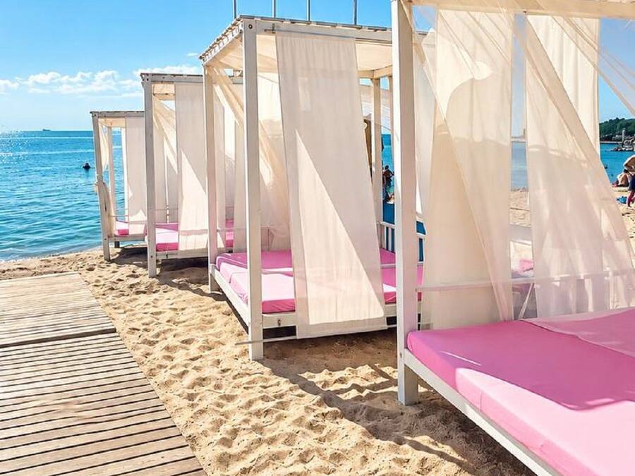 A row of beach cabanas with white canopies and pink cushions, set on a wooden deck by the sea.
