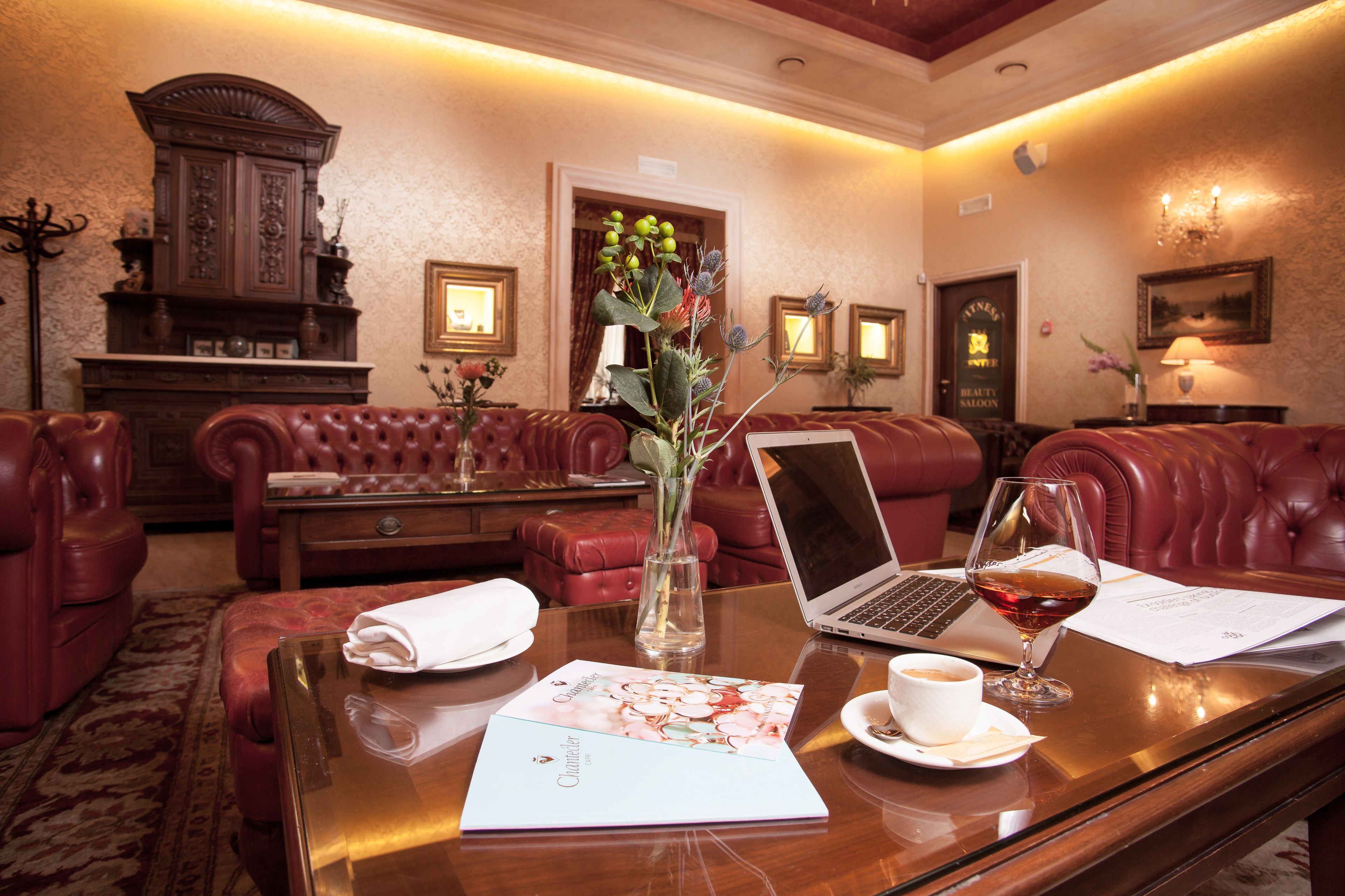 A hotel lobby with leather seating, a wooden cabinet, a laptop, a glass of red wine, and a vase with flowers.