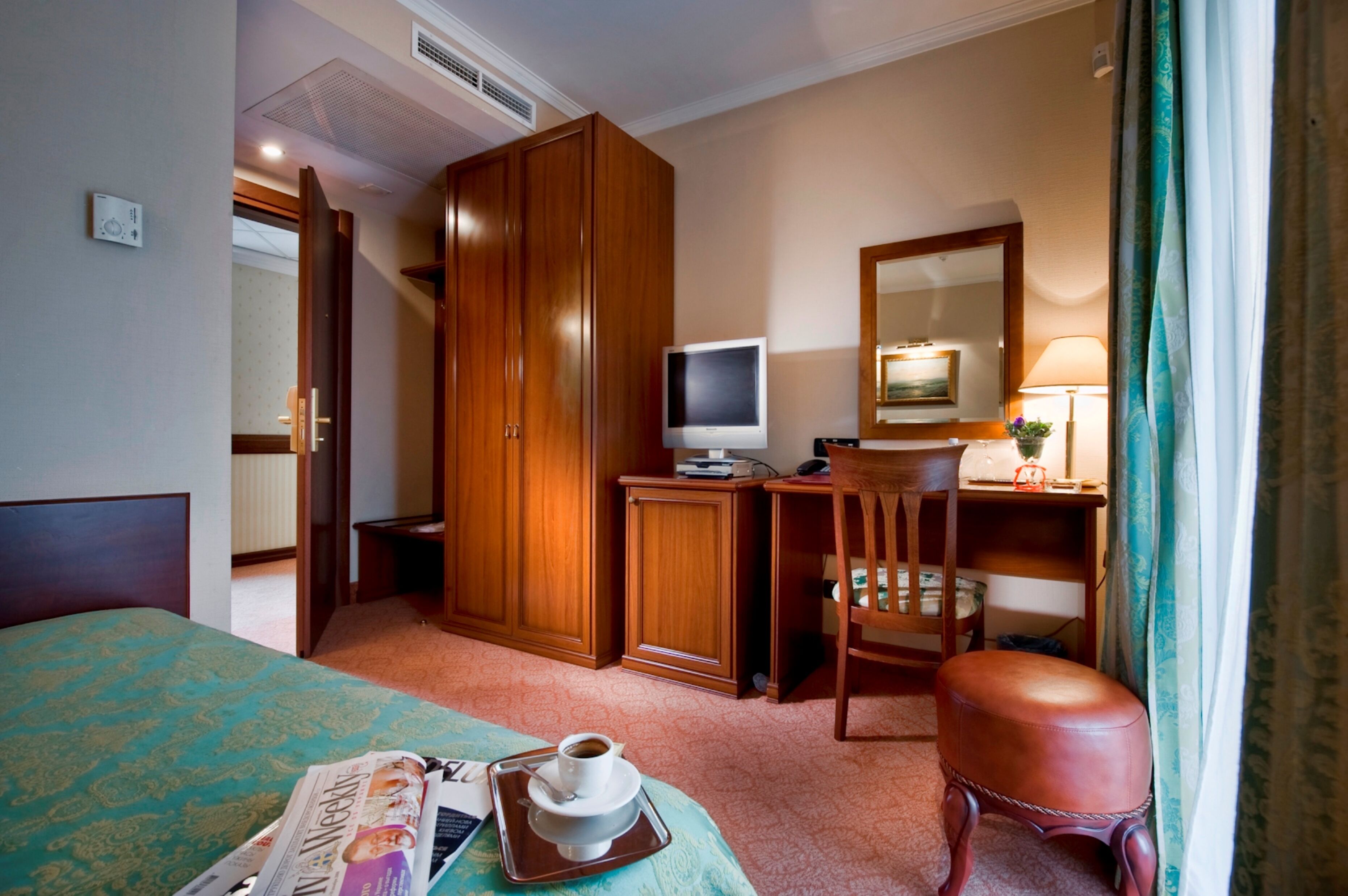 Une chambre d’hôtel avec un lit, un bureau avec un ordinateur, une chaise, une armoire et une petite table sur laquelle se trouvent une tasse de café et un journal.