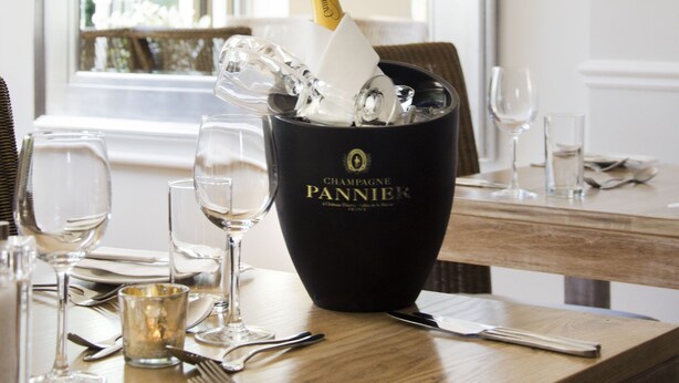 Mesa de comedor decorada con una botella de champán, copas y cubiertos, con un espejo grande que refleja la vista de una ventana.