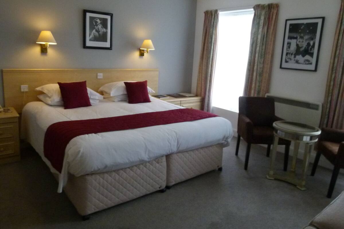 Habitación de hotel con una cama, dos lámparas de noche, una silla, una mesita y dos cuadros en la pared.