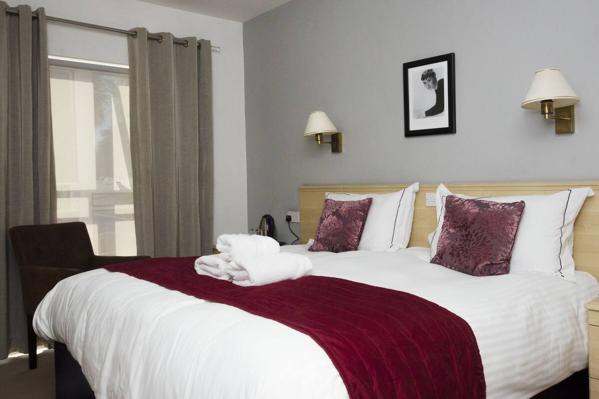 Una habitación de hotel con cama, sillón, escritorio y ventana con cortinas.