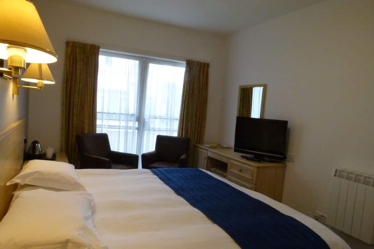 Una habitación de hotel con una cama, dos sillas, un escritorio con televisor y un ventanal con cortinas.