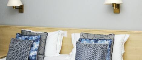 Cama bem arrumada com cobertor cinza, almofadas com estampas geométricas e duas luminárias fixadas na parede cinza-clara.