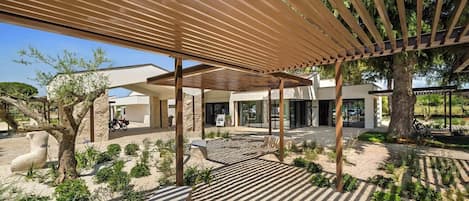 Ein überdachter Außenbereich mit einer Pergola, ein modernes Gebäude und eine sandige Landschaft mit spärlicher Vegetation.