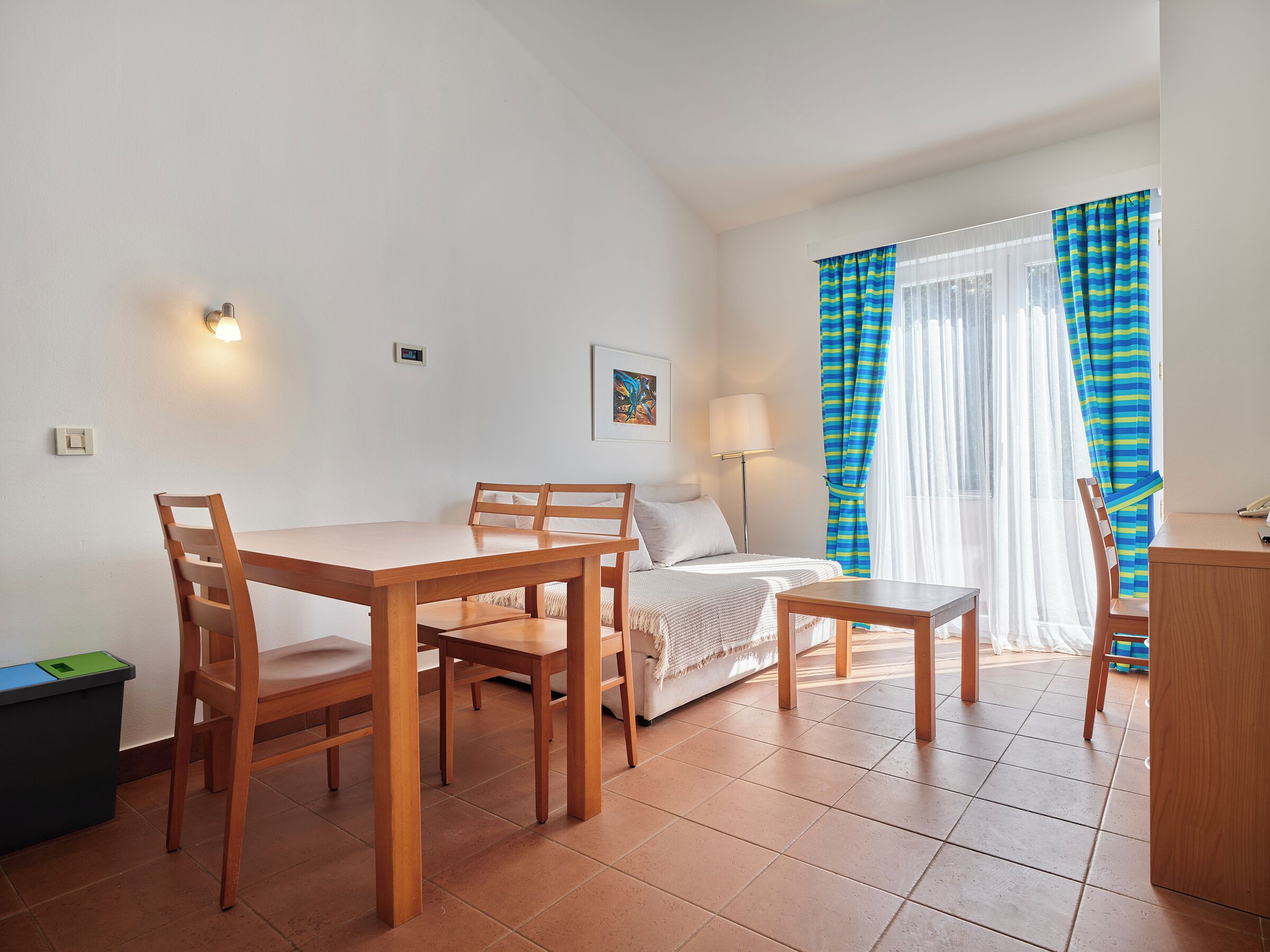 A small, bright room with a bed, a table, and chairs. There is a window with curtains, a lamp, and a picture on the wall.