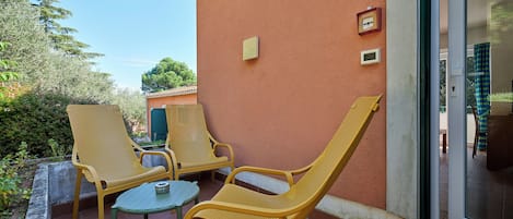 A patio with yellow wicker chairs and a small table, a door with a number sign, and a view of greenery.