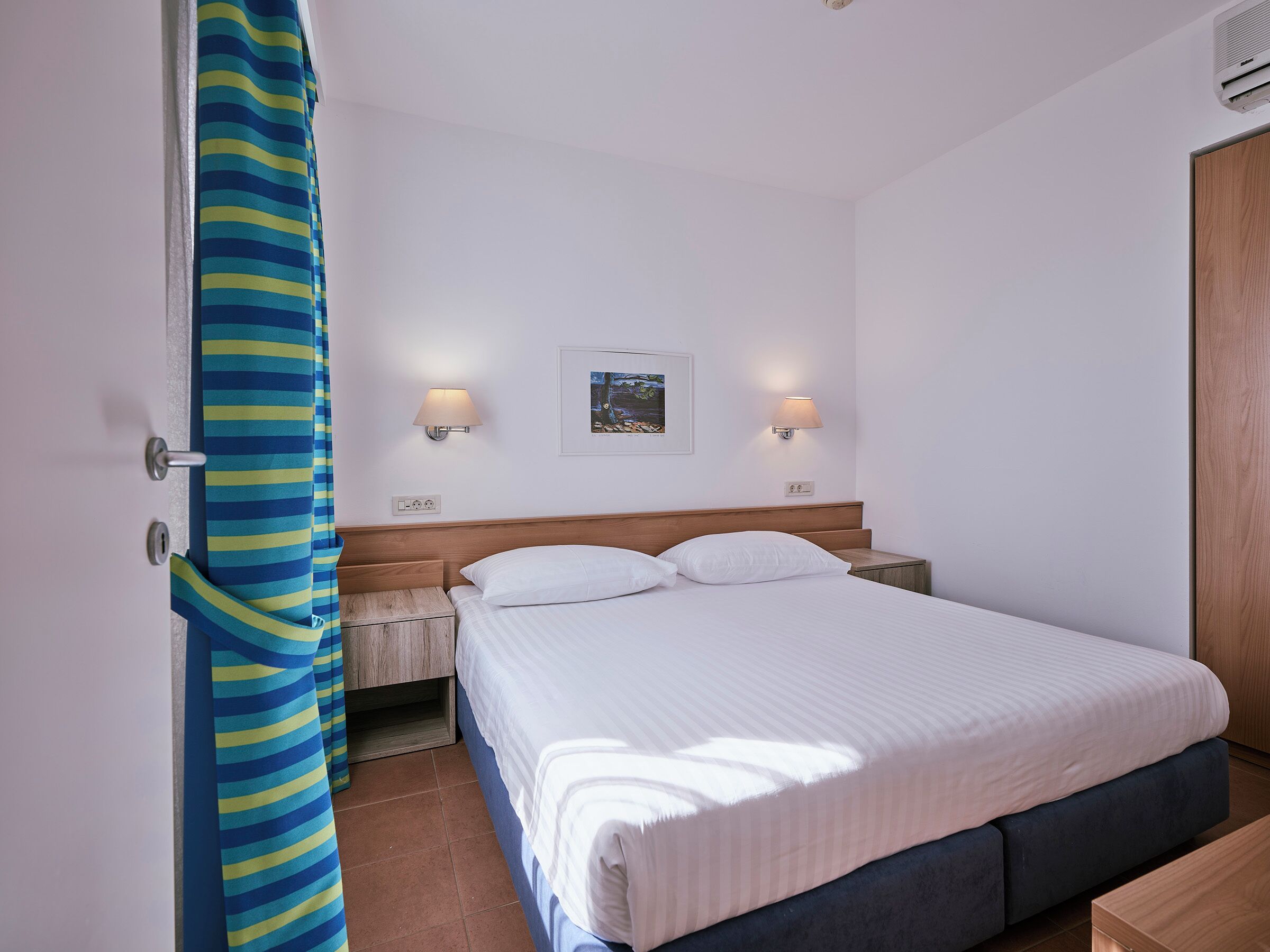 A hotel room with a bed, two bedside lamps, a nightstand, a wooden headboard, and a wall-mounted picture.