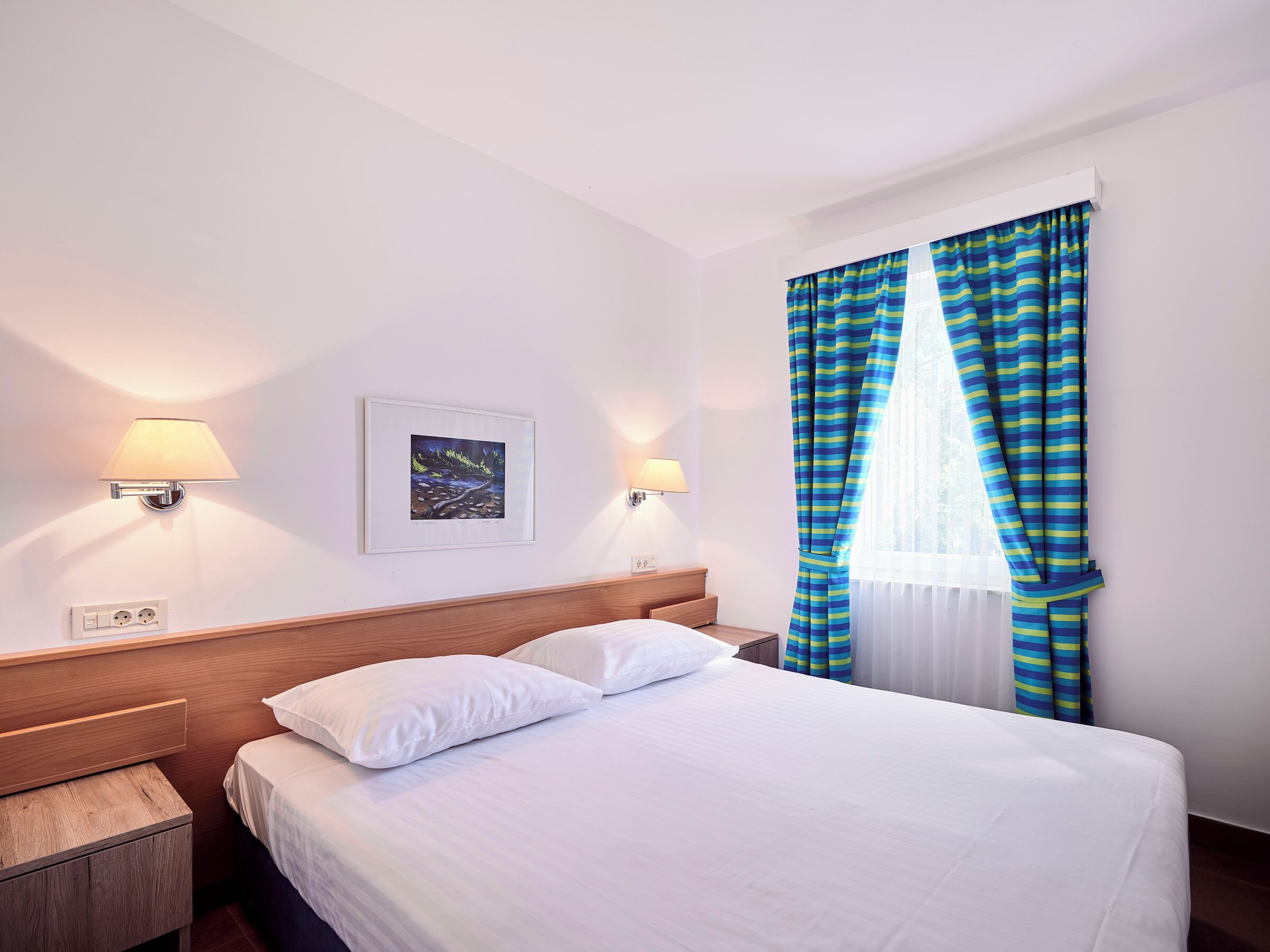 A hotel room with a bed, two bedside lamps, a framed picture on the wall, and striped curtains.