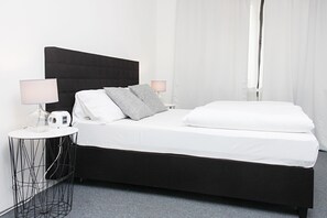 Una habitación de hotel moderna con cabecera negra, ropa de cama blanca y una mesita de noche pequeña con una lámpara.