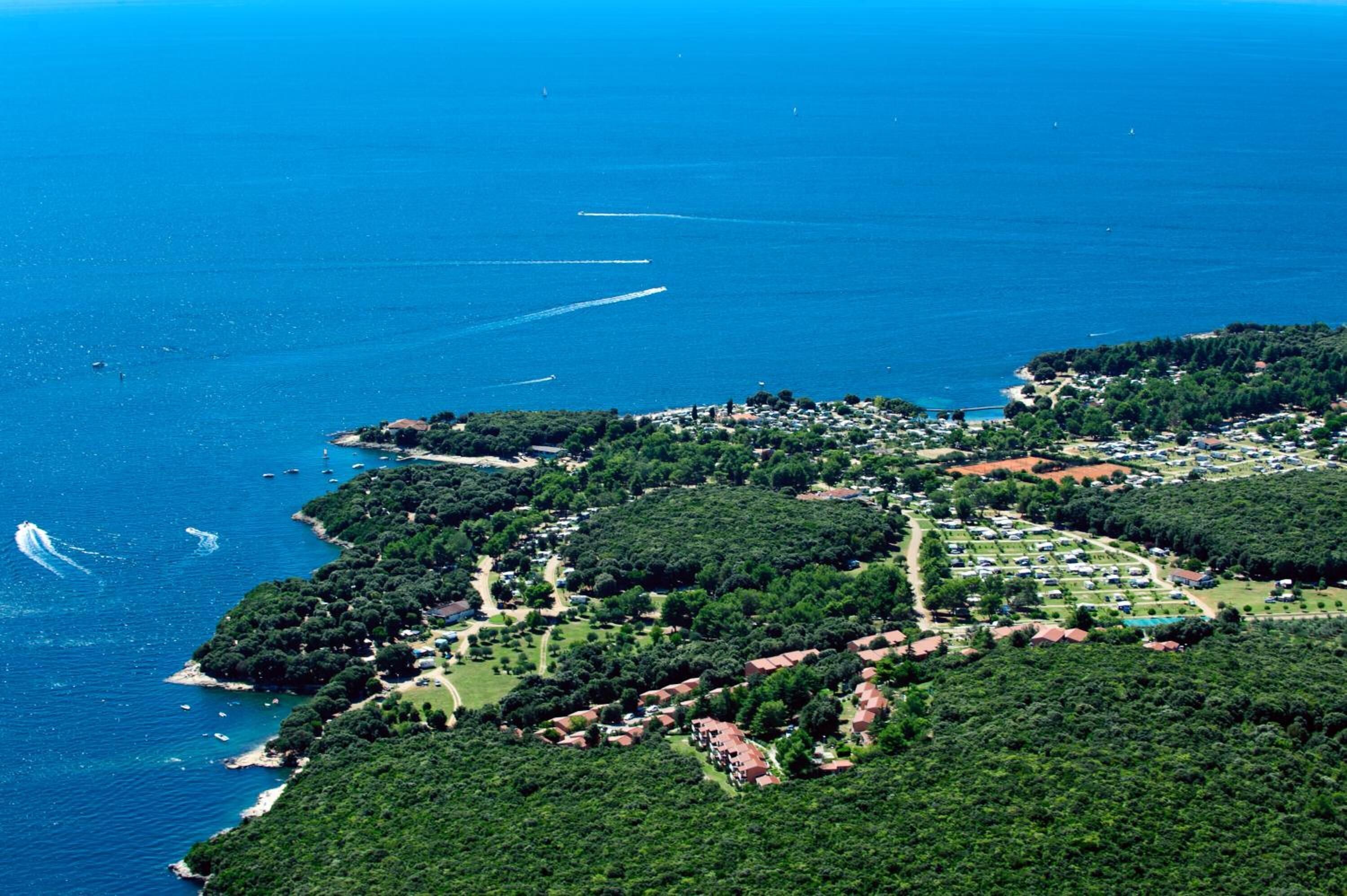 Luchtbeeld van een kustplaatsje met weelderige begroeiing, een jachthaven met boten en een heldere blauwe zee.