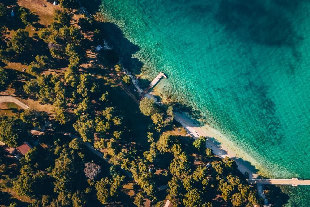 Ett flygbildsbild av ett kustområde med en pir som sträcker sig ut i det klara, turkosfärgade vattnet, omgivet av tät vegetation och några få byggnader.