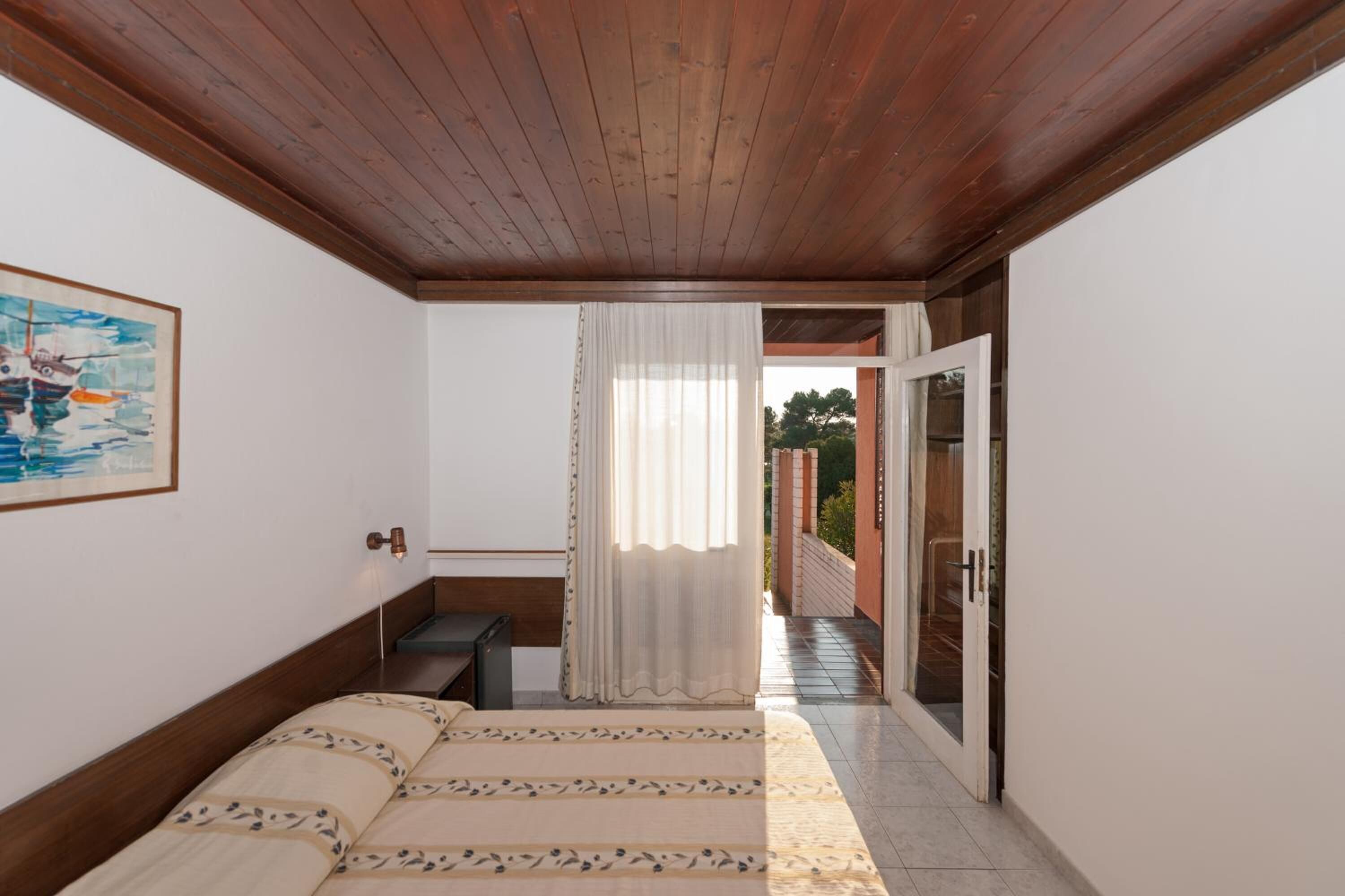 A bedroom with a wooden ceiling, a bed with a patterned bedspread, a nightstand, a painting on the wall, and a view through a door to an outdoor area.