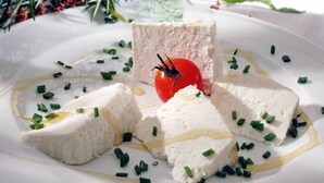 Un plato blanco con un trozo de comida blanca, posiblemente tofu, decorado con cebollines verdes y un tomate rojo.