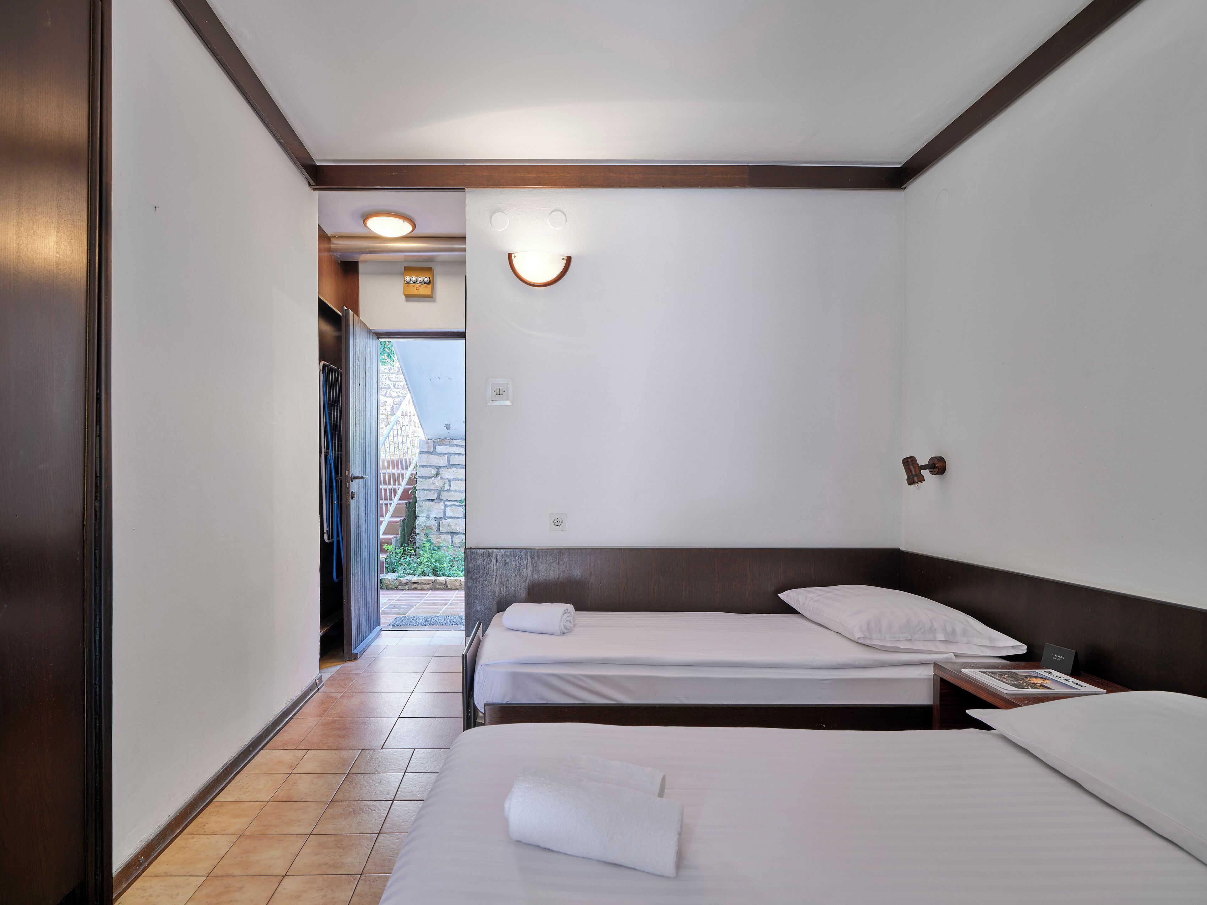 A hotel room with two beds, a wooden headboard, a small bedside table with a magazine, and a view of a stone wall and greenery outside.