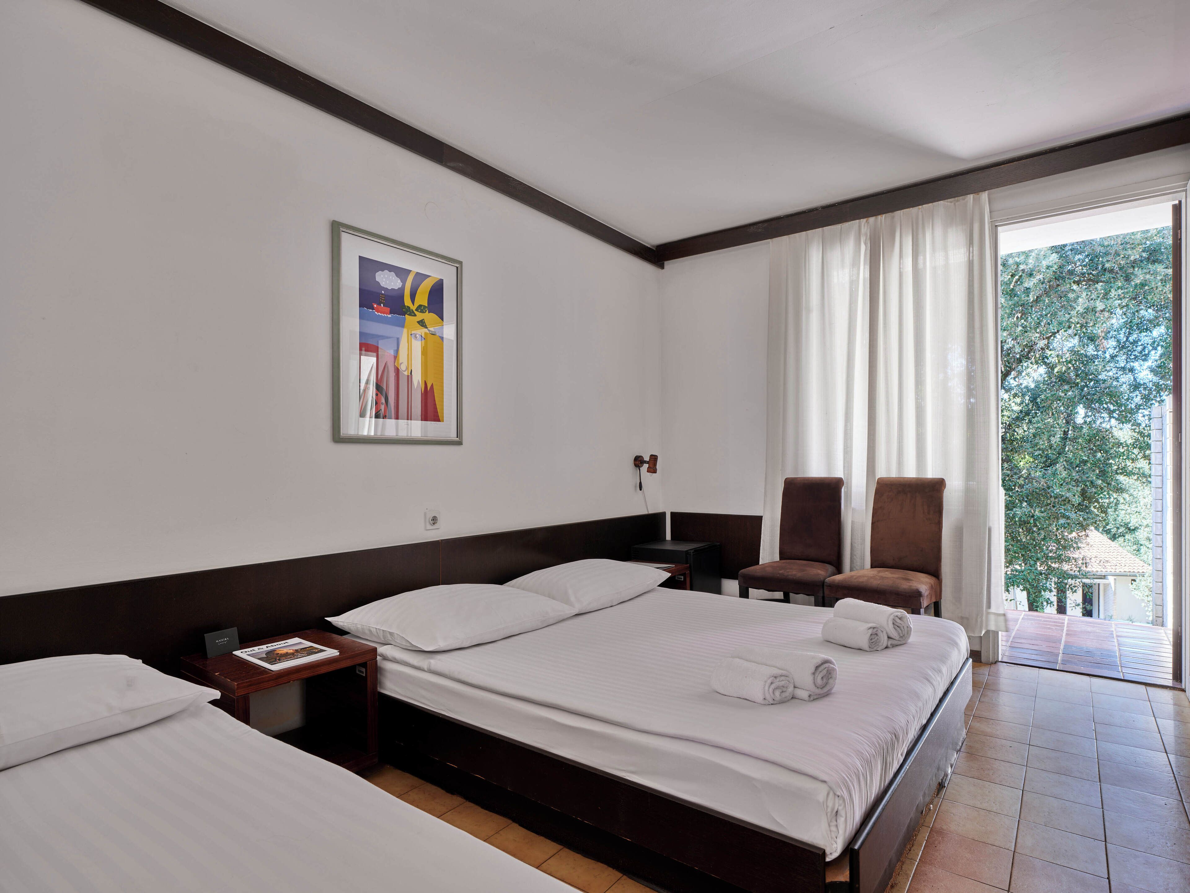 Quarto de hotel com uma cama grande, duas cadeiras, uma mesa de cabeceira e um quadro na parede.