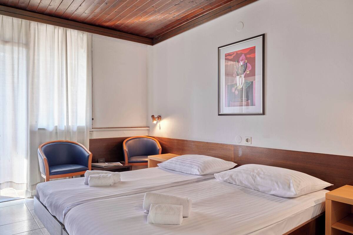 Une chambre d’hôtel avec un plafond en bois, un lit avec du linge de lit blanc, deux chaises, une table de chevet et un tableau encadré au mur.
