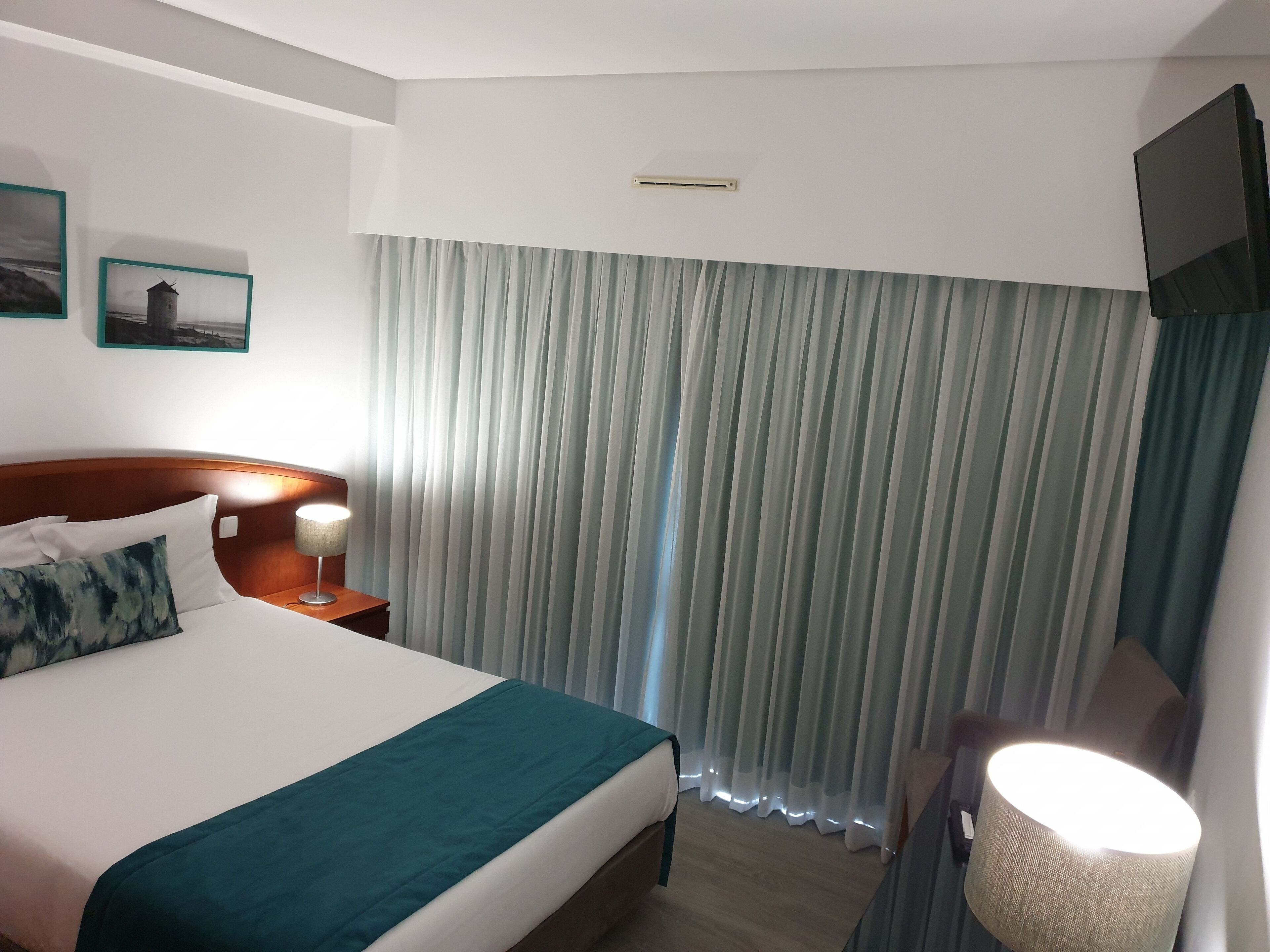 A hotel room with a bed, a nightstand with a lamp, a TV mounted on the wall, and a window with curtains.