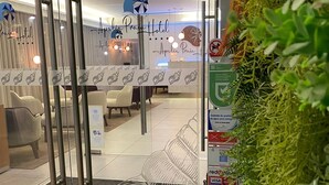 Un espace d’accueil d’hôtel avec une porte en verre, un comptoir de réception et un coin salon avec des chaises. Des panneaux indiquent les équipements et les services disponibles, et un mur végétal agrémente l’espace.