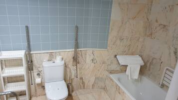 A bathroom with a toilet, a handrail, a shelf unit, and a bathtub.
