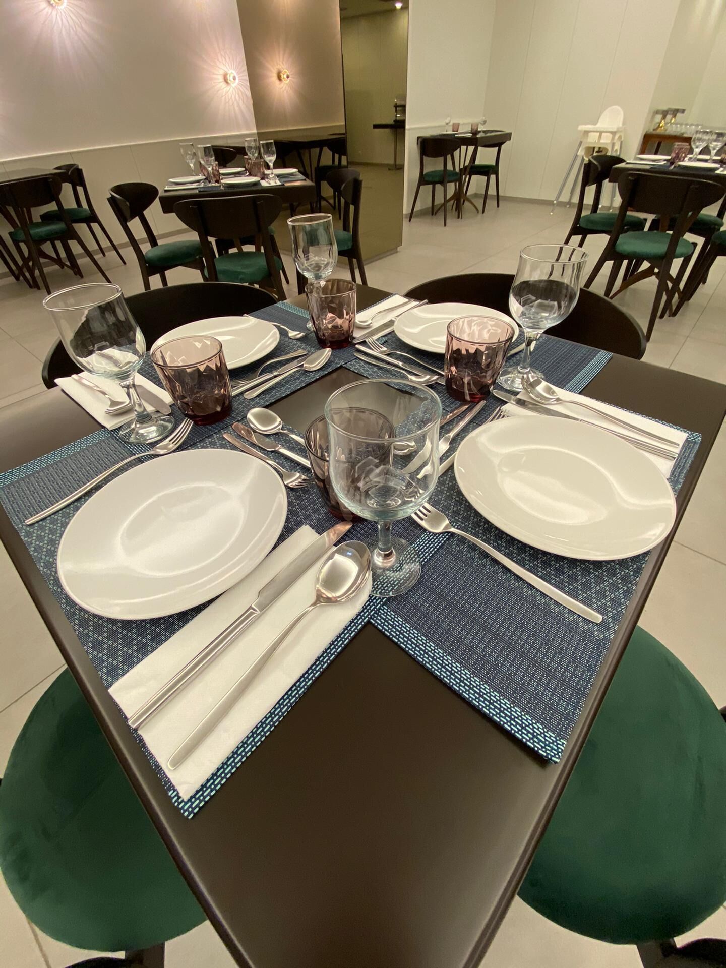 Una mesa de comedor dispuesta con platos blancos, cubiertos y vasos transparentes, rodeada de sillas tapizadas de verde.
