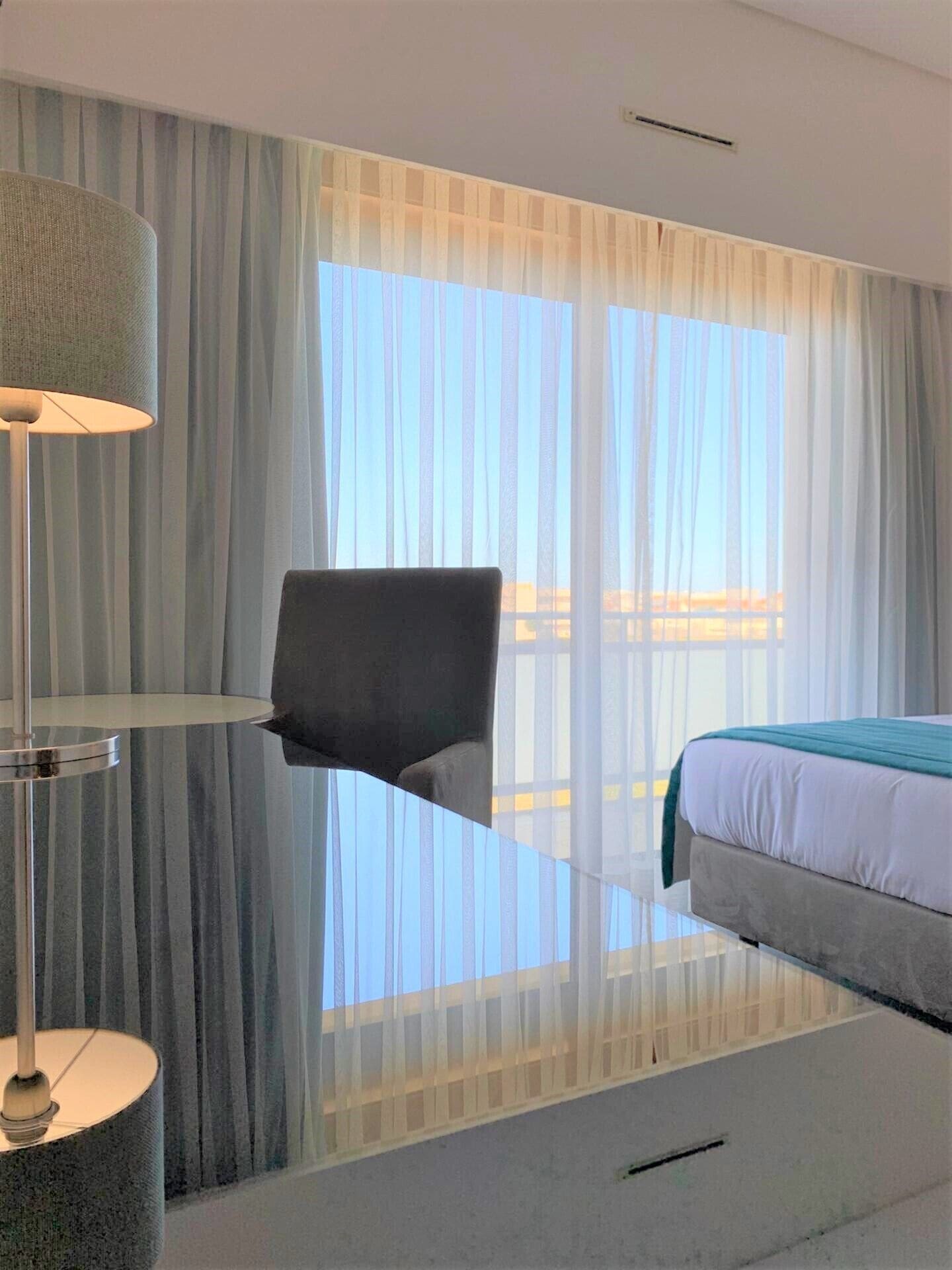 Ett hotellrum med en säng, en stol, en lampa och tunna, genomskinliga gardiner.