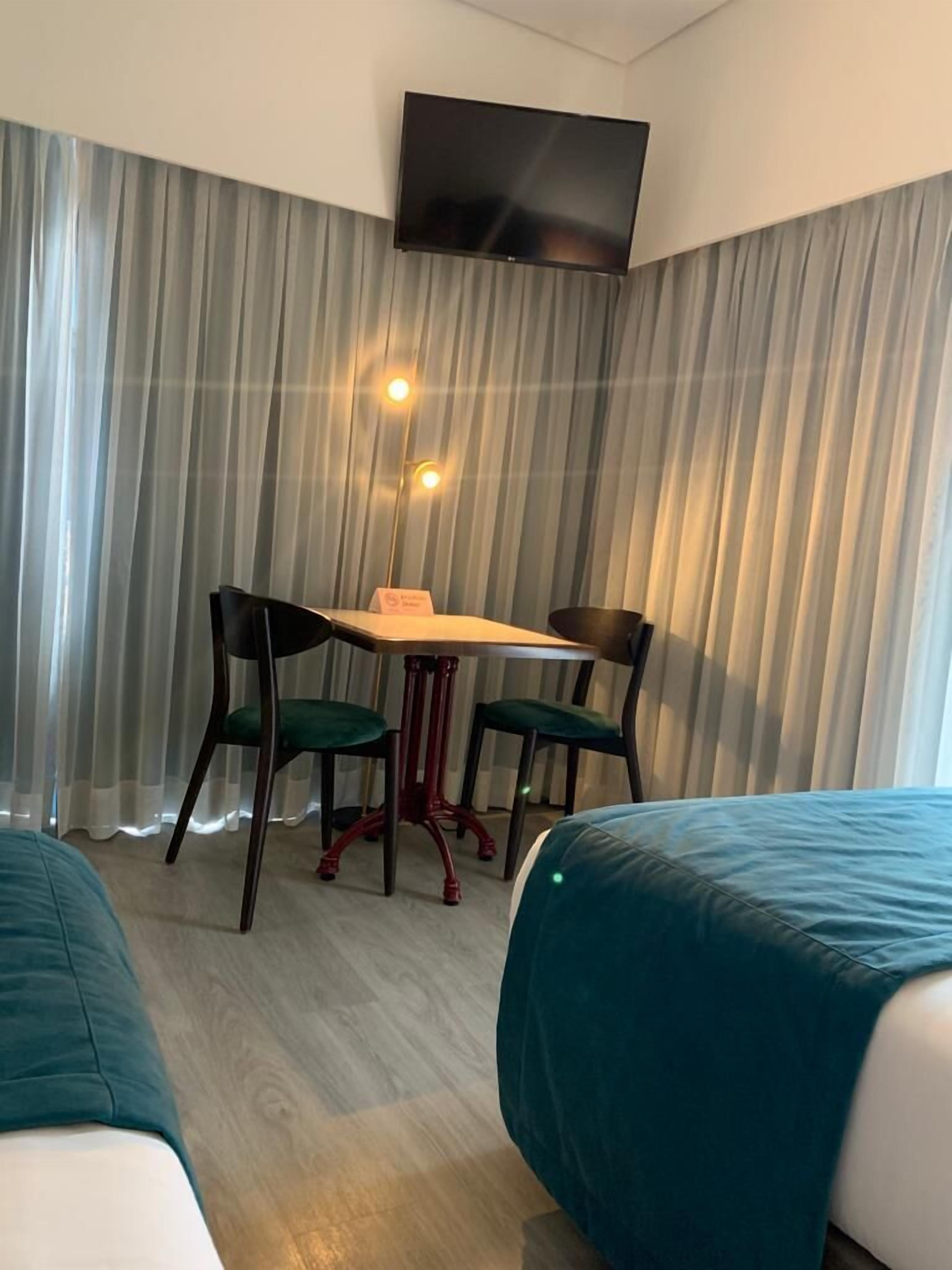Une chambre d’hôtel avec deux lits, un bureau avec des chaises, un téléviseur à écran plat fixé au mur et des rideaux légers.
