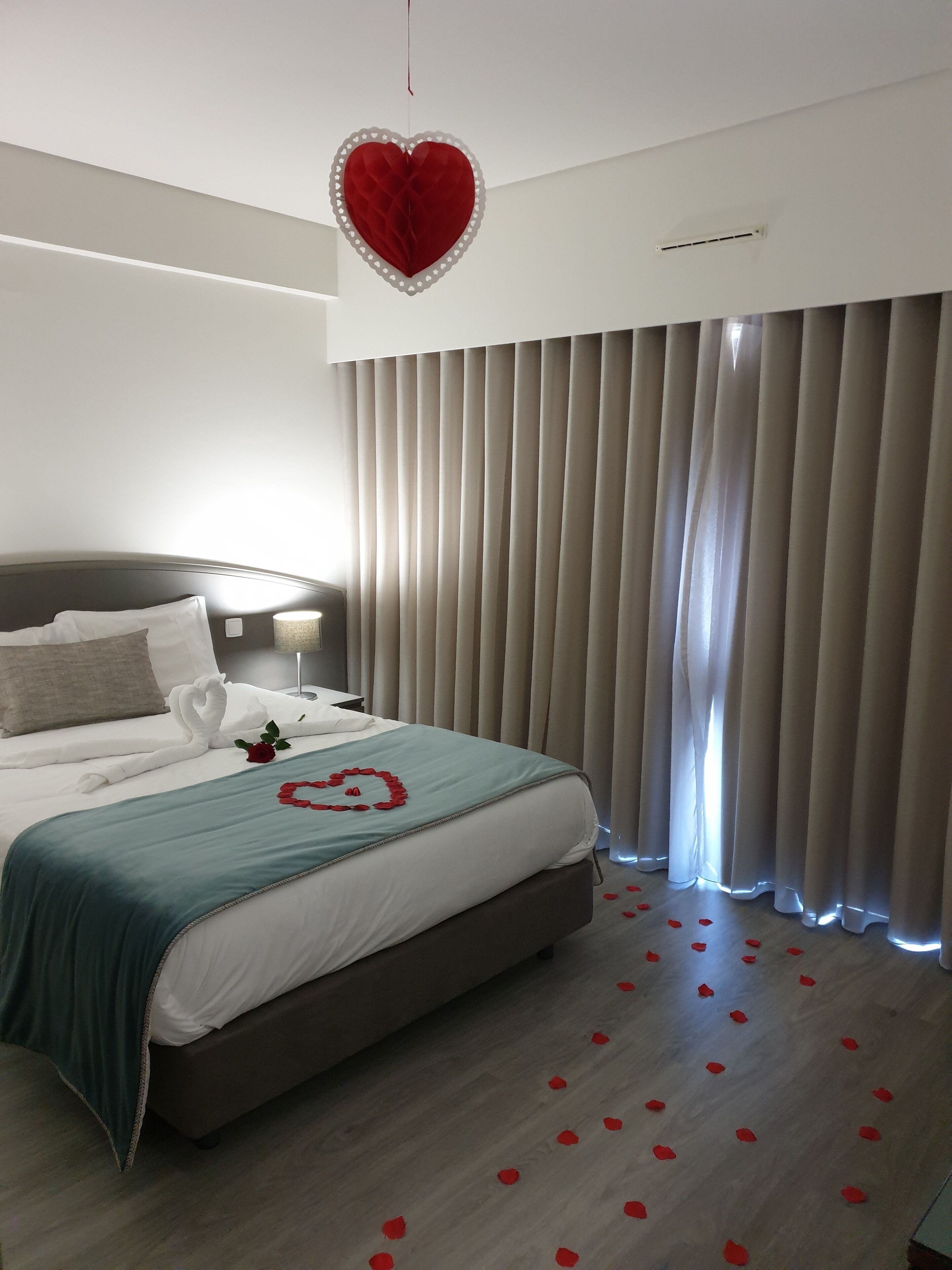 Une chambre d’hôtel moderne avec un lit soigneusement fait, une décoration en forme de cœur sur le lit et des pétales de rose éparpillés sur le sol.