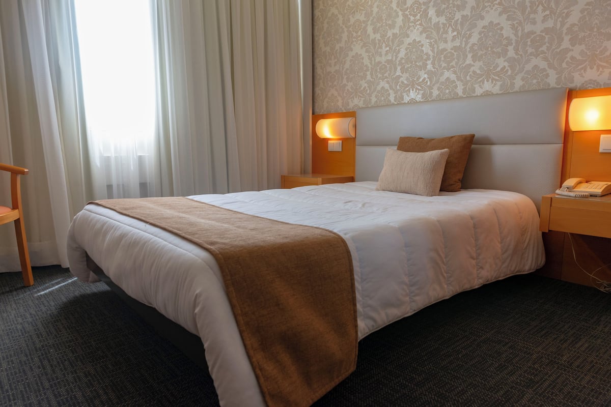 A hotel room with a large bed, a wooden nightstand with a telephone, and a wall-mounted lamp.
