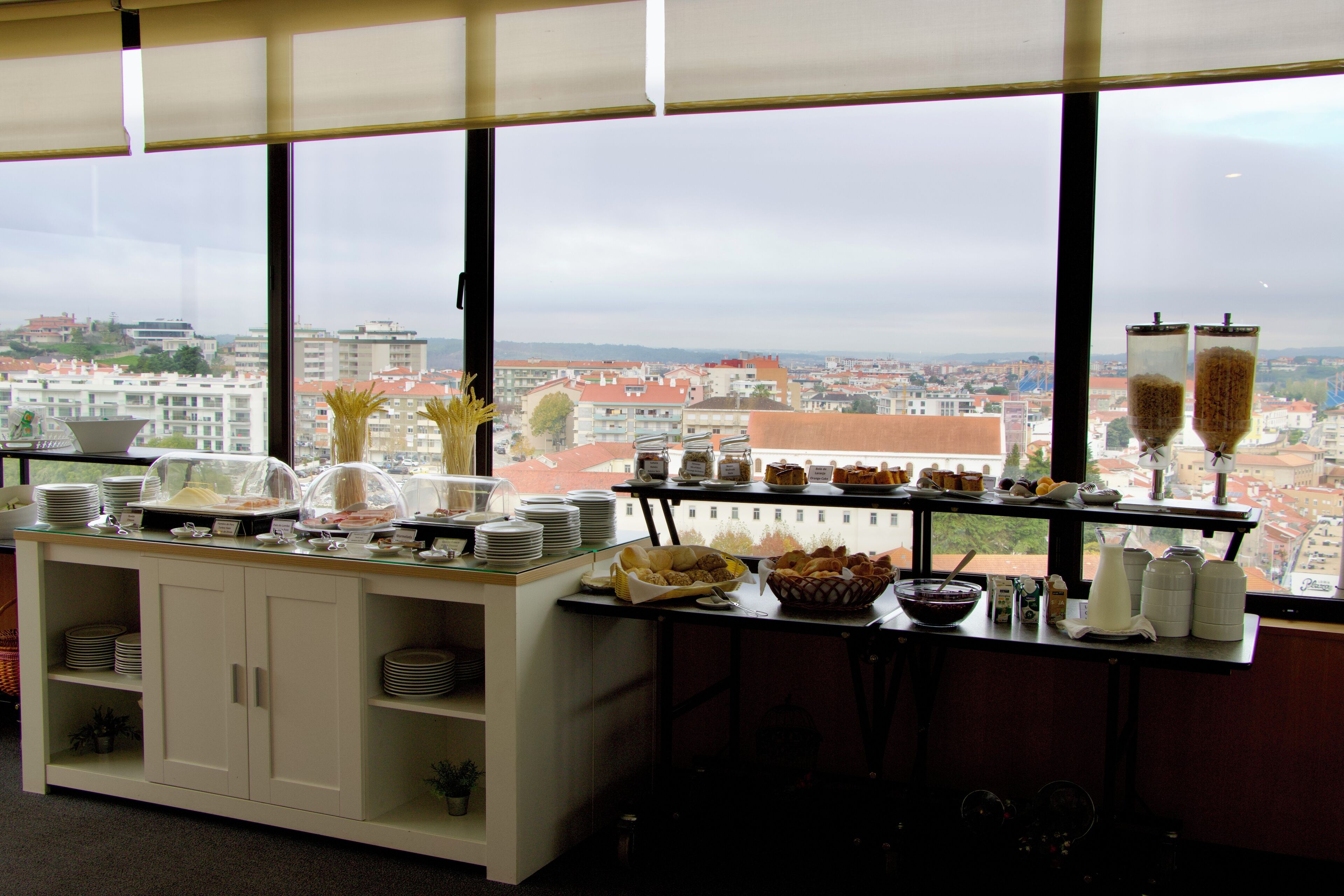 Montaje de bufé con diferentes platillos y vista panorámica de la ciudad a través de amplias ventanas.