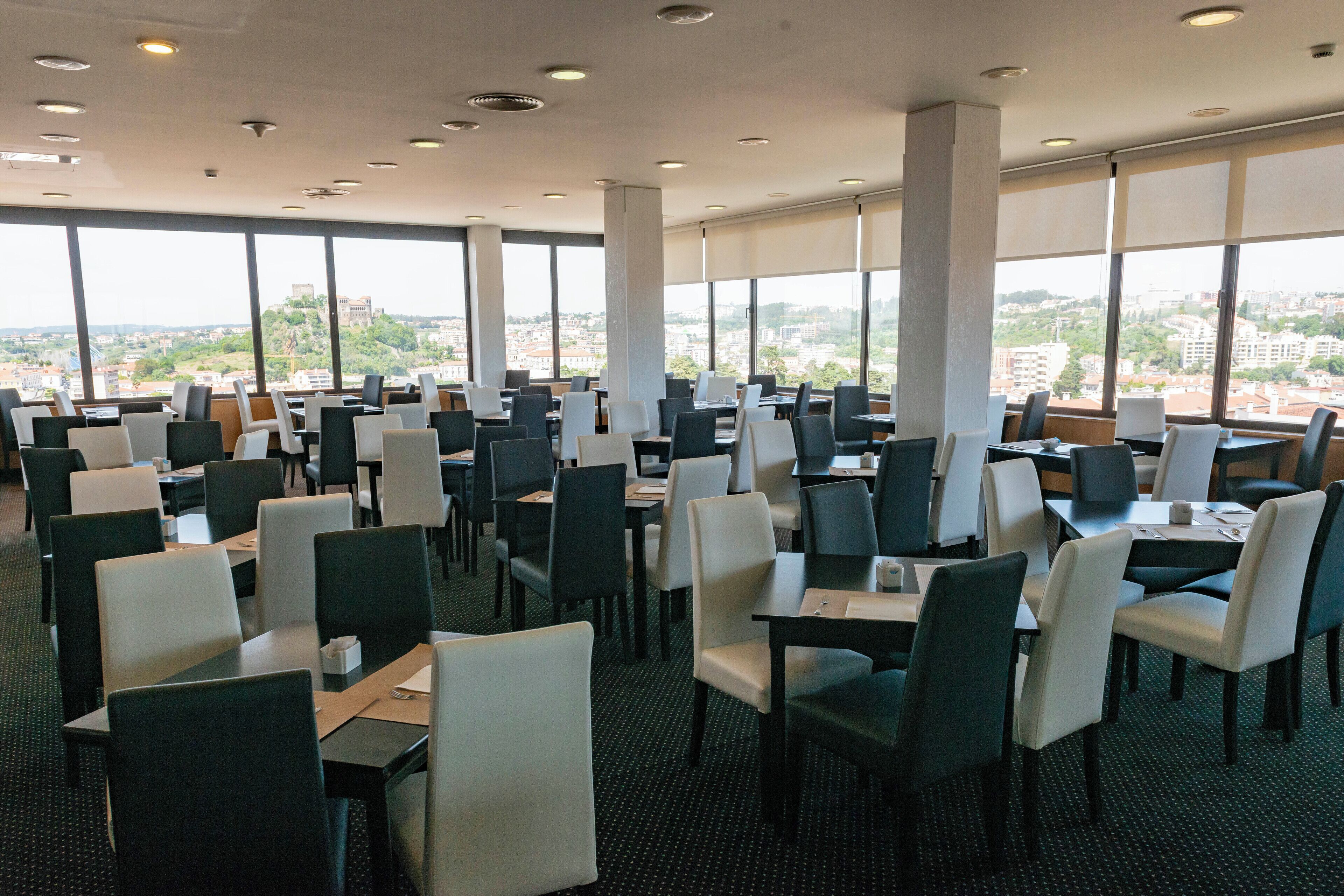A spacious dining area with multiple tables and chairs, large windows offering a city view, and a high ceiling with recessed lighting.
