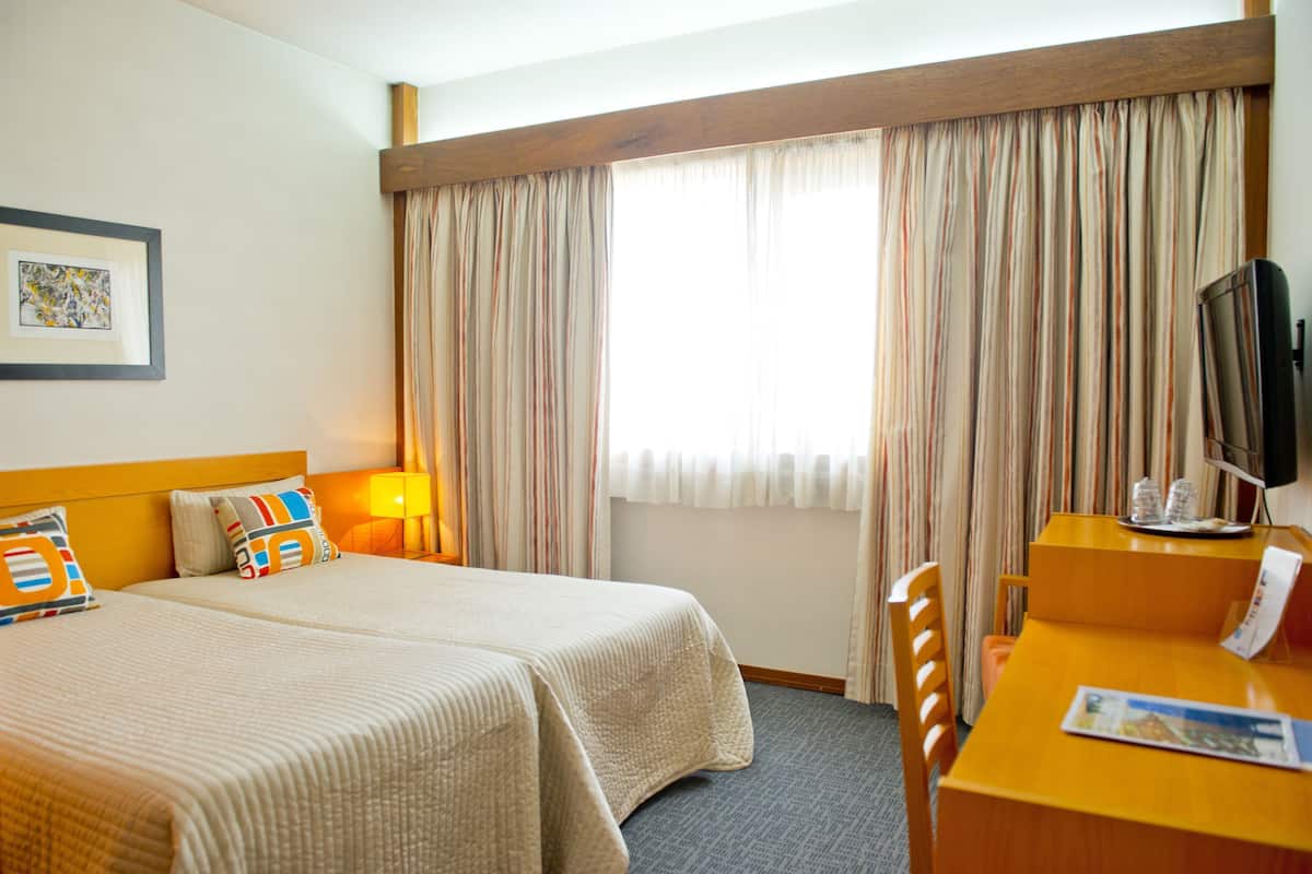 A hotel room with two beds, a desk, a television, and a window with curtains.