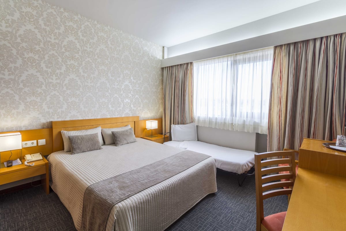 A hotel room with a large bed, a smaller bed, a desk, and a chair. There are two bedside tables with lamps and a telephone.